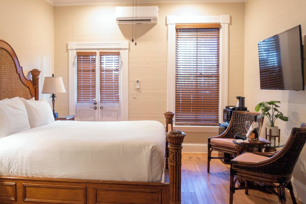 A bedroom with a large bed, two windows with blinds, a television on the wall, and a chair with a table.