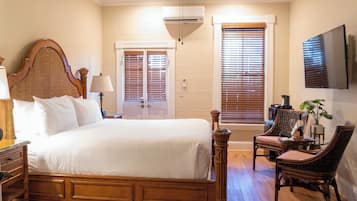 A bedroom with a large bed, two windows with blinds, a television on the wall, and a chair with a table.