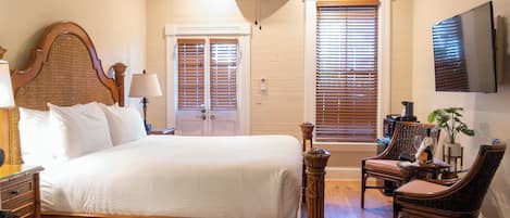 A bedroom with a large bed, two windows with blinds, a television on the wall, and a chair with a table.