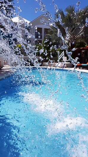 A swimming pool with water splashing, surrounded by palm trees and a building in the background.