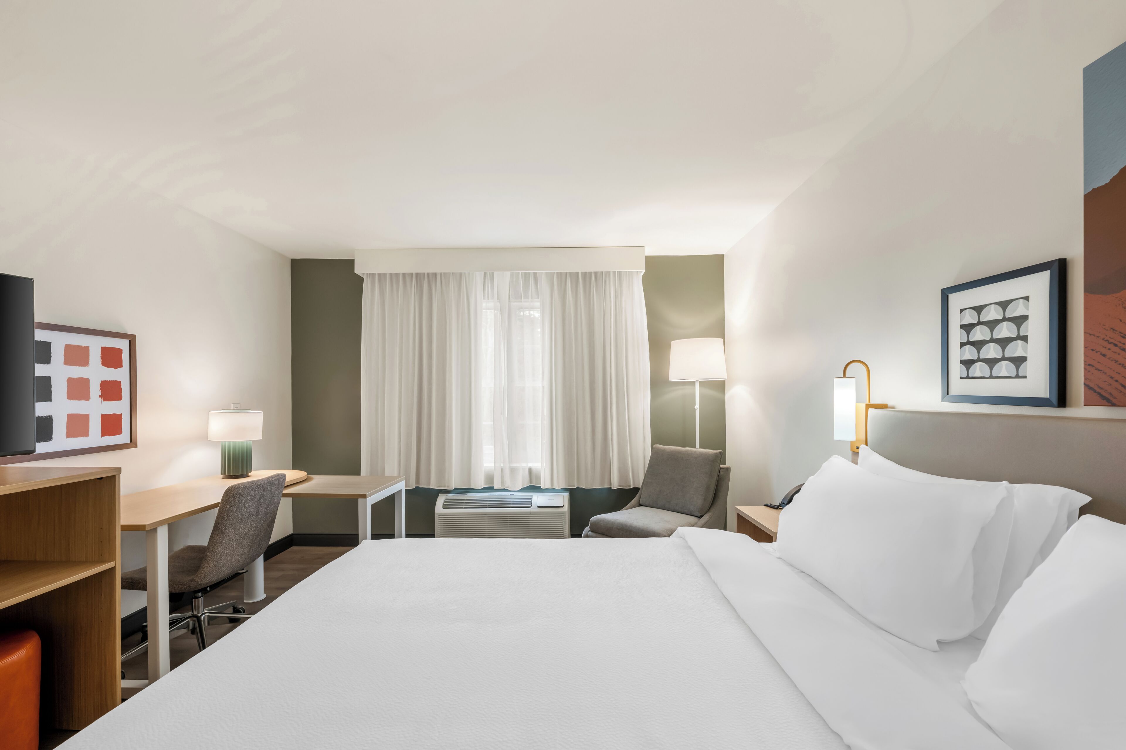 A hotel room with a bed, a desk, a chair, a lamp, and a window with curtains.