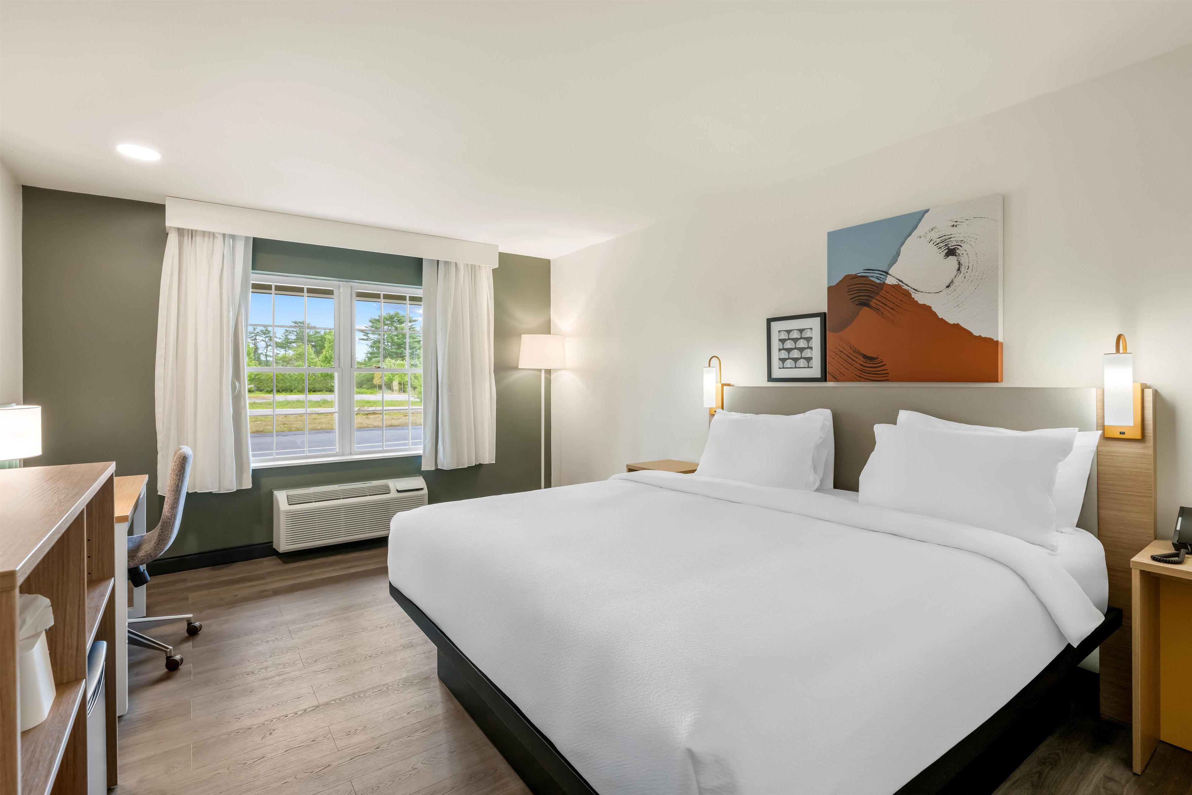 A hotel room with a bed, desk, chair, and a view of greenery through the window.