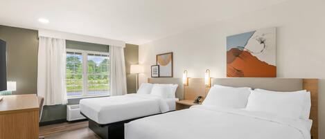 A hotel room with two beds, a desk, a chair, a window with curtains, a lamp, a framed picture, and a wall-mounted artwork.