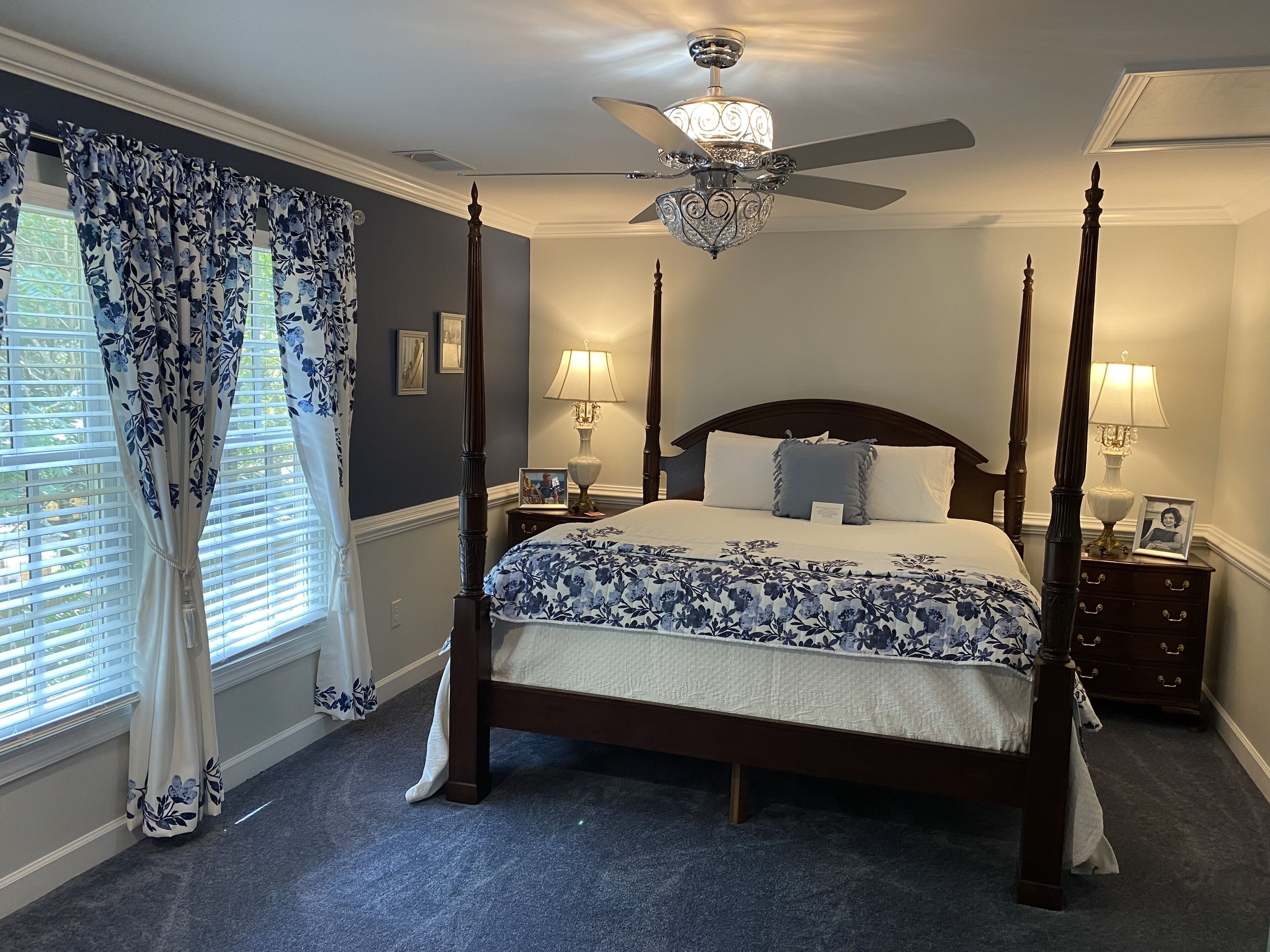A four-poster bed with a chandelier ceiling fan, a nightstand with a lamp, and a window with curtains.