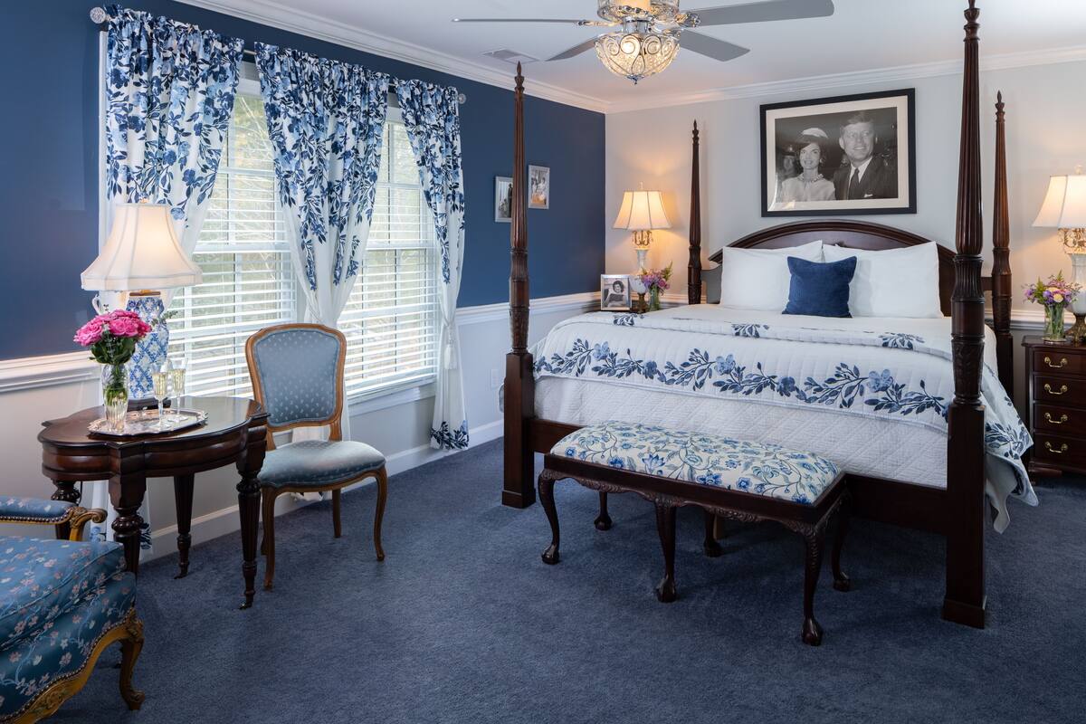 Un dormitorio con una cama con dosel, una silla, una mesa con un florero y una ventana con cortinas azules.