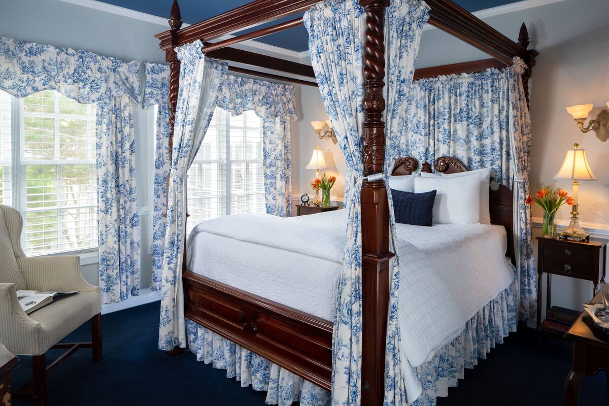Una cama con dosel y cortinas florales, una mesita de noche con lámpara y una silla con un libro.