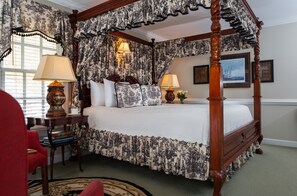 Una cama con dosel, con colcha estampada de flores, dos lámparas de noche, un sillón rojo y un cuadro enmarcado en la pared.