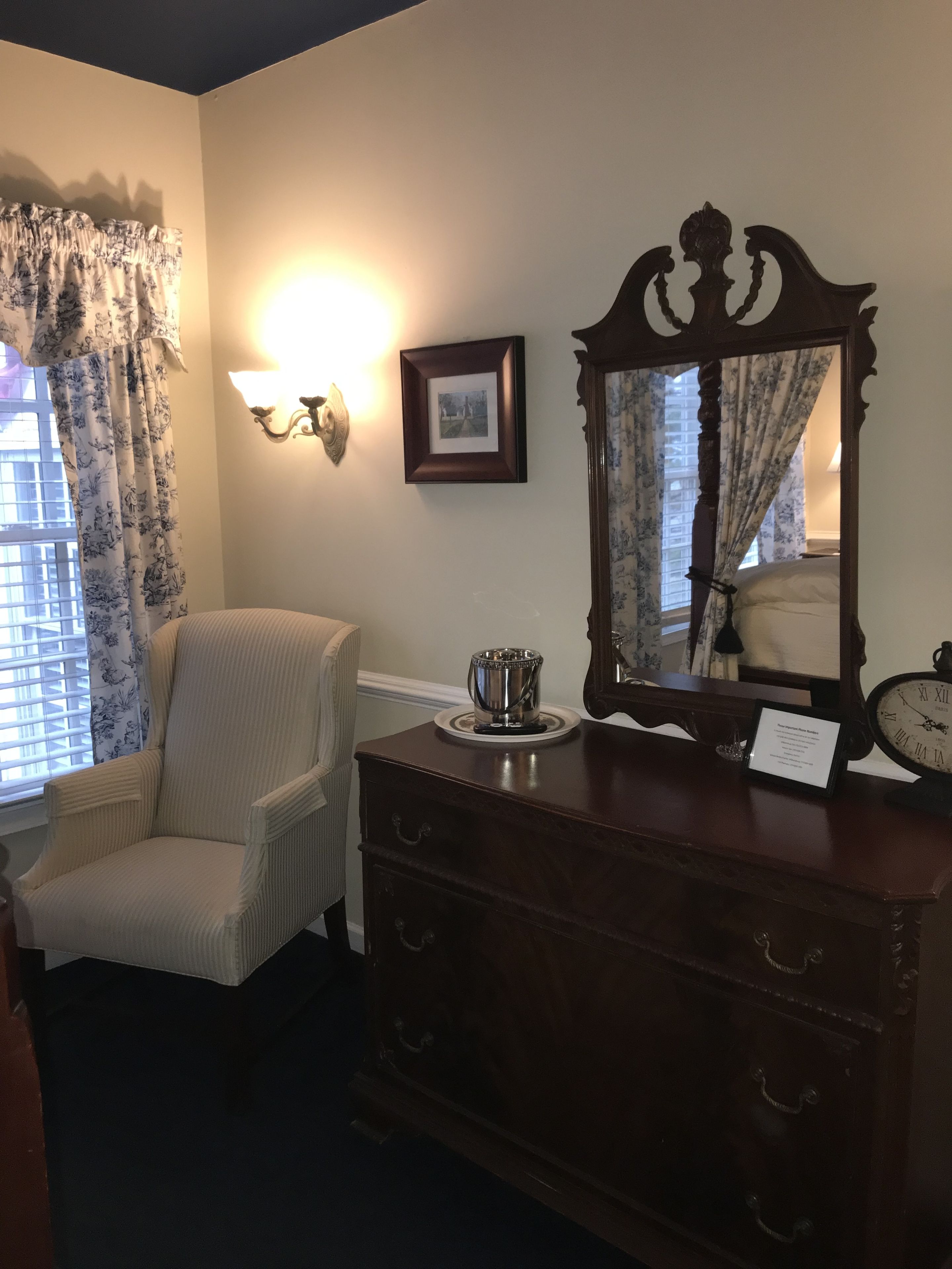 A room with a wooden dresser, a framed picture on the wall, a chair, and a window with floral curtains.