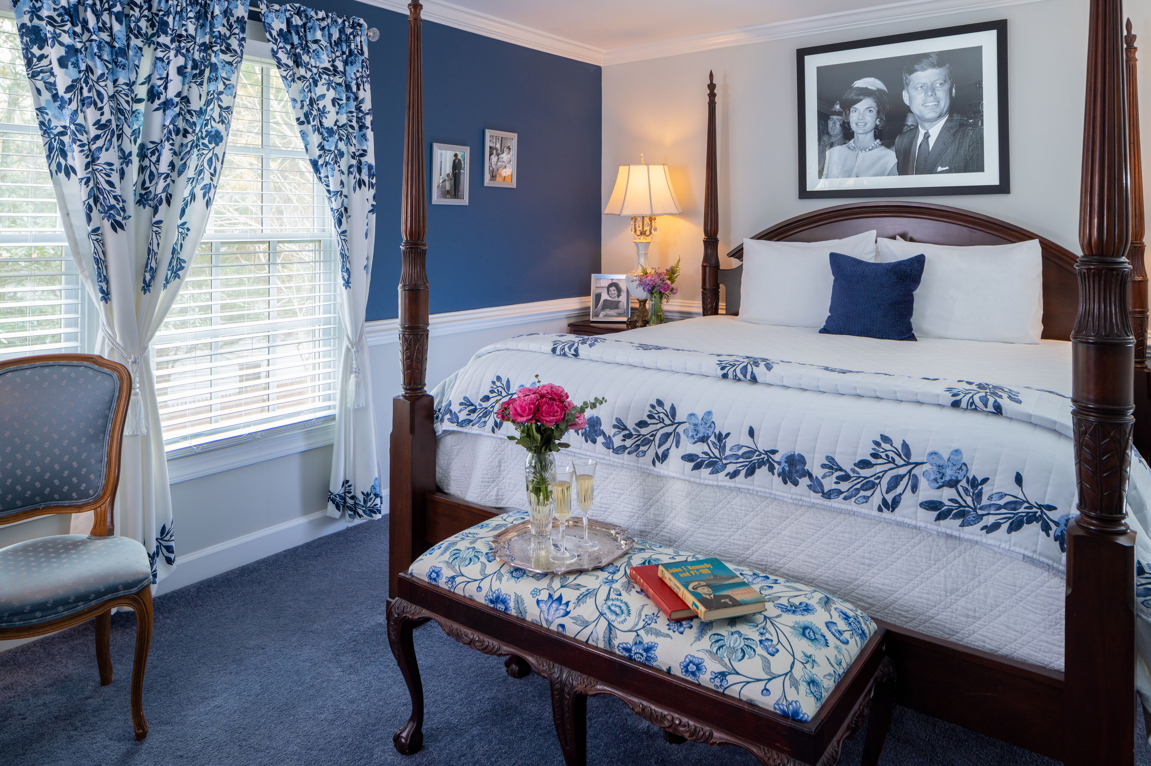 A four-poster bed with a floral bedspread, a bedside table with a lamp, a chair, and a tray with a vase of flowers and glasses.