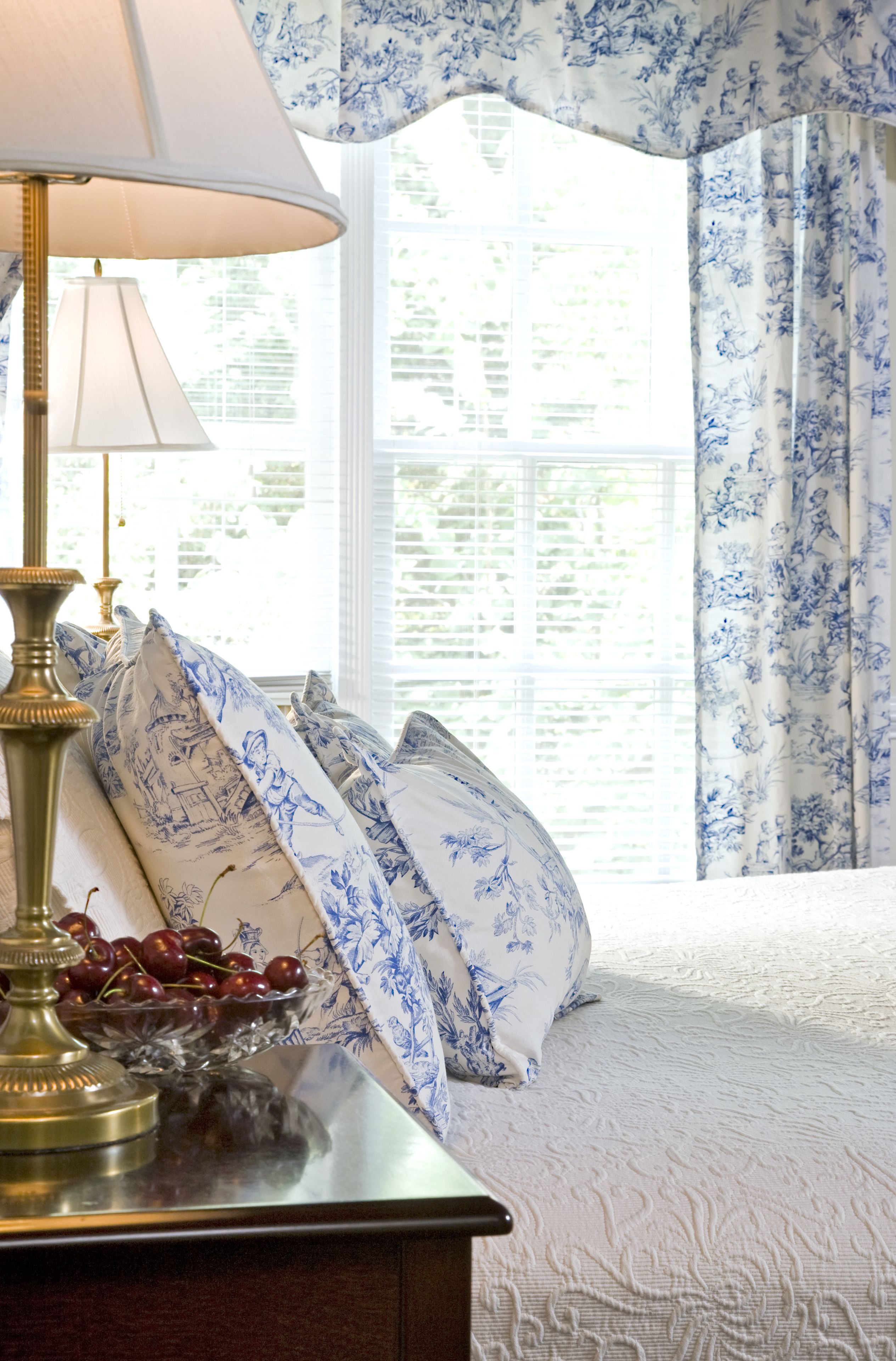 A neatly made bed with a white and blue floral bedspread, a brass lamp, and a bowl of cherries on a wooden nightstand.