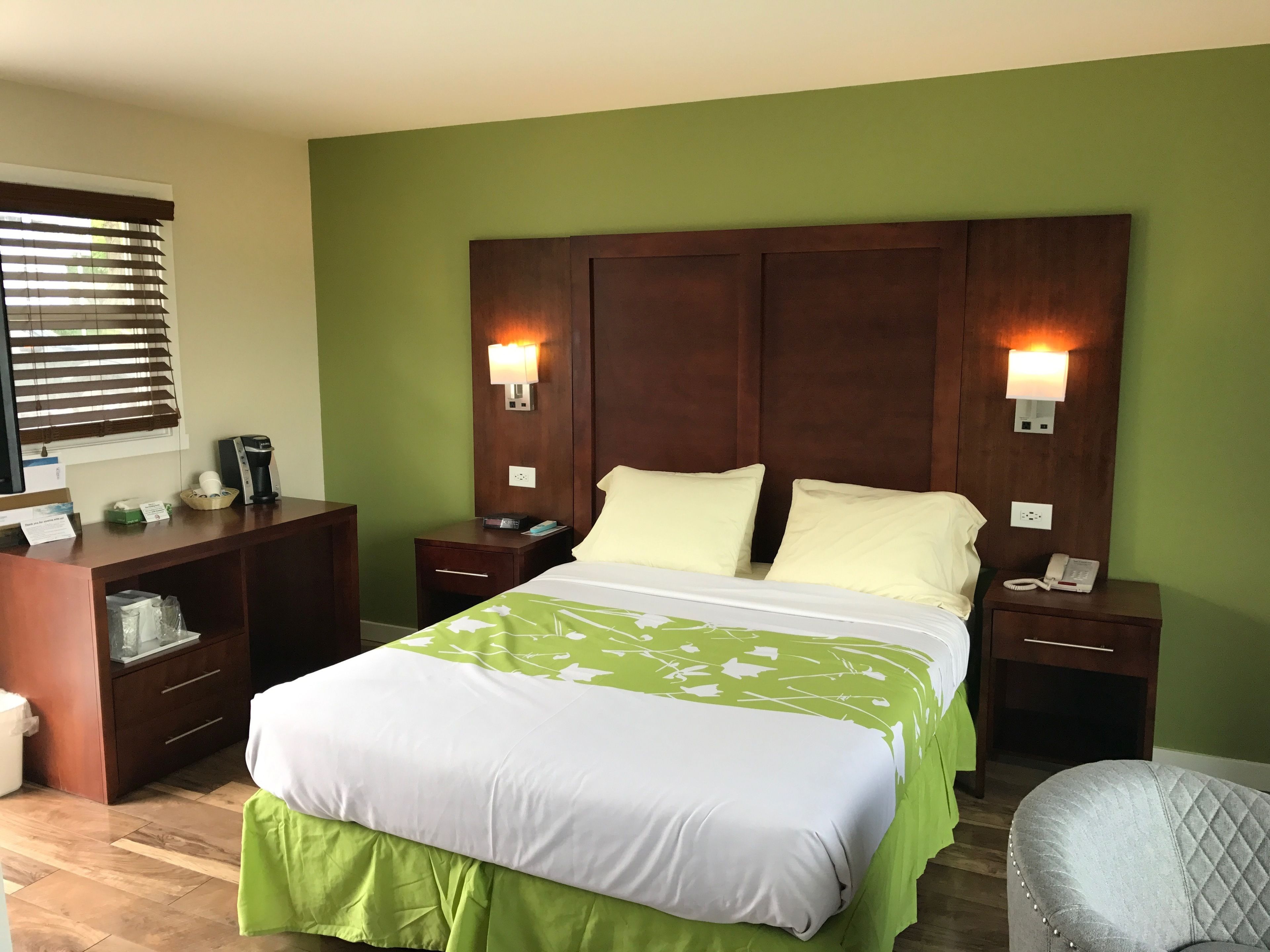 Habitación de hotel con una cama grande, un escritorio con cafetera, dos lámparas de noche y una silla.