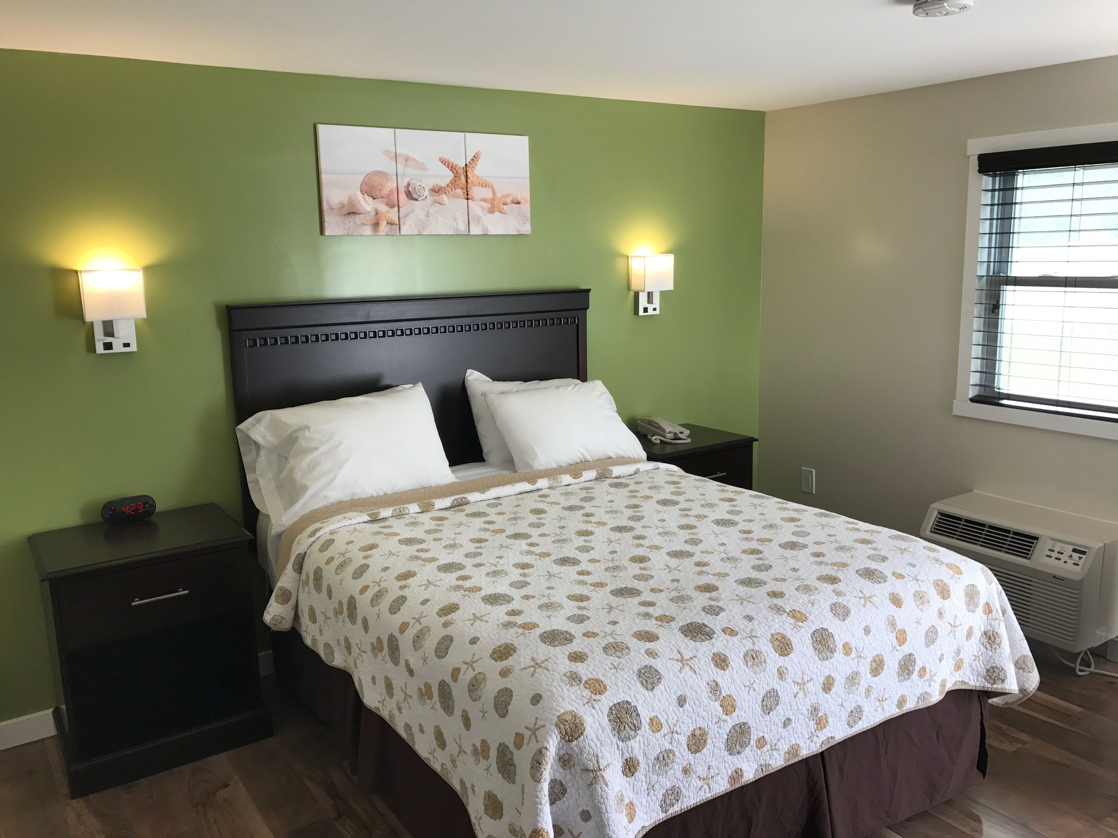 Habitación de hotel con cama, dos mesitas de noche, lámpara de pared, una obra de arte en la pared que representa criaturas marinas, y ventana con persianas.