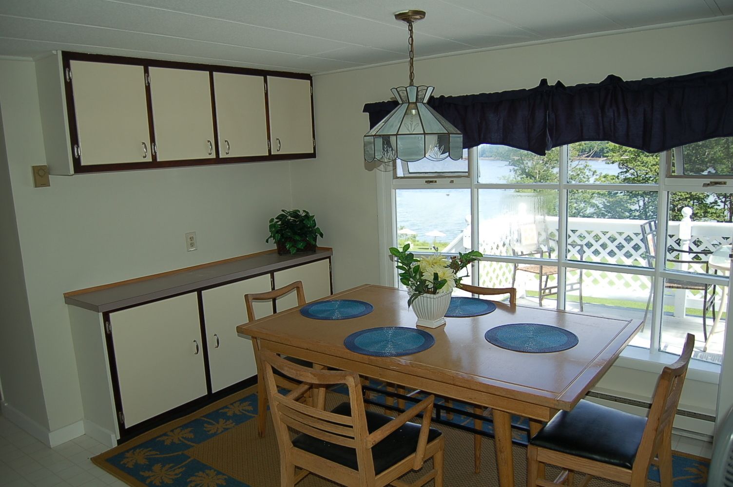 A dining area with a wooden table, blue placemats, and chairs. There is a window with a view of the outdoors, a ceiling light fixture, and a plant on the table.