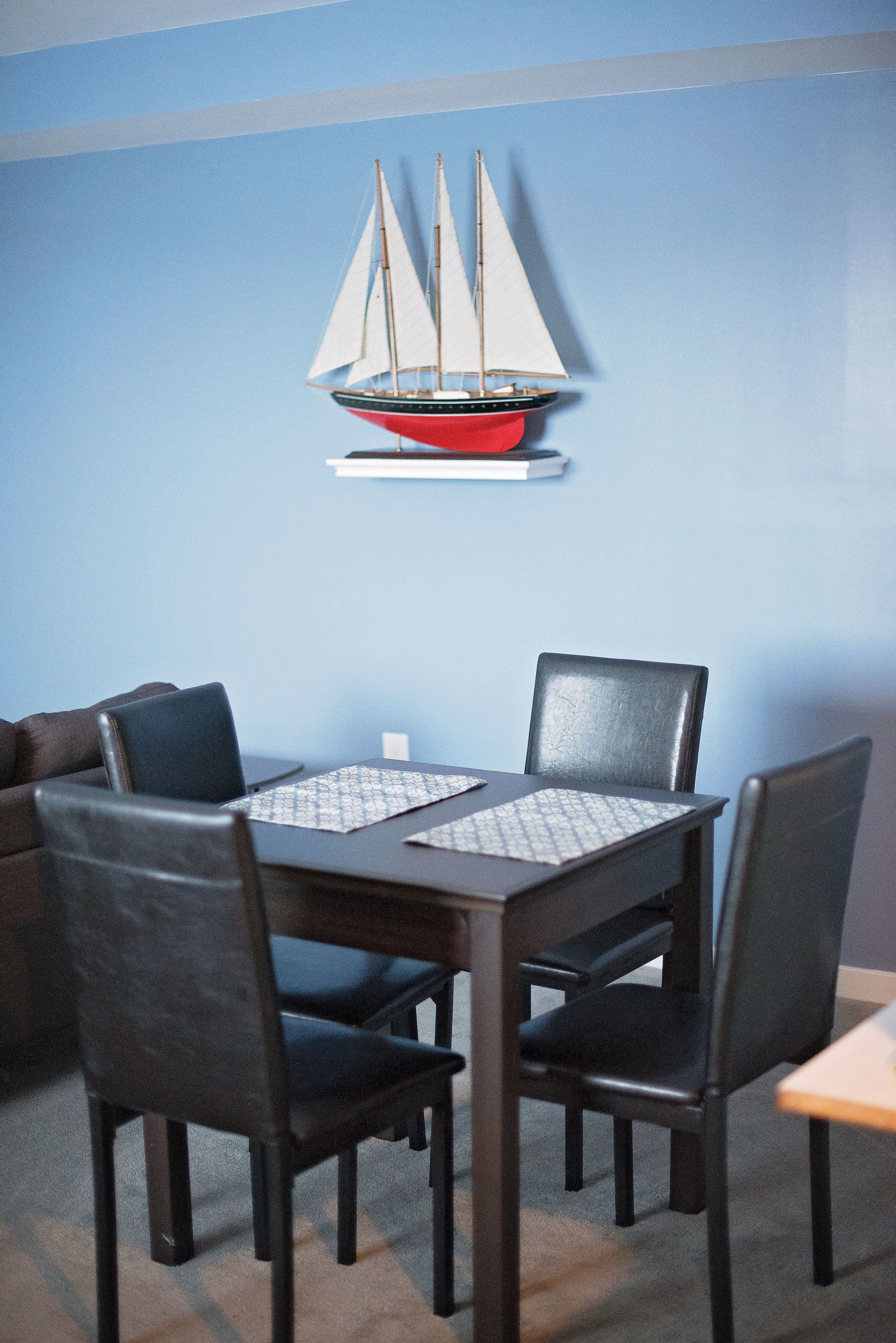 Uma área de refeições com uma mesa e quatro cadeiras, uma pequena decoração de barco à vela na parede e uma parede azul.