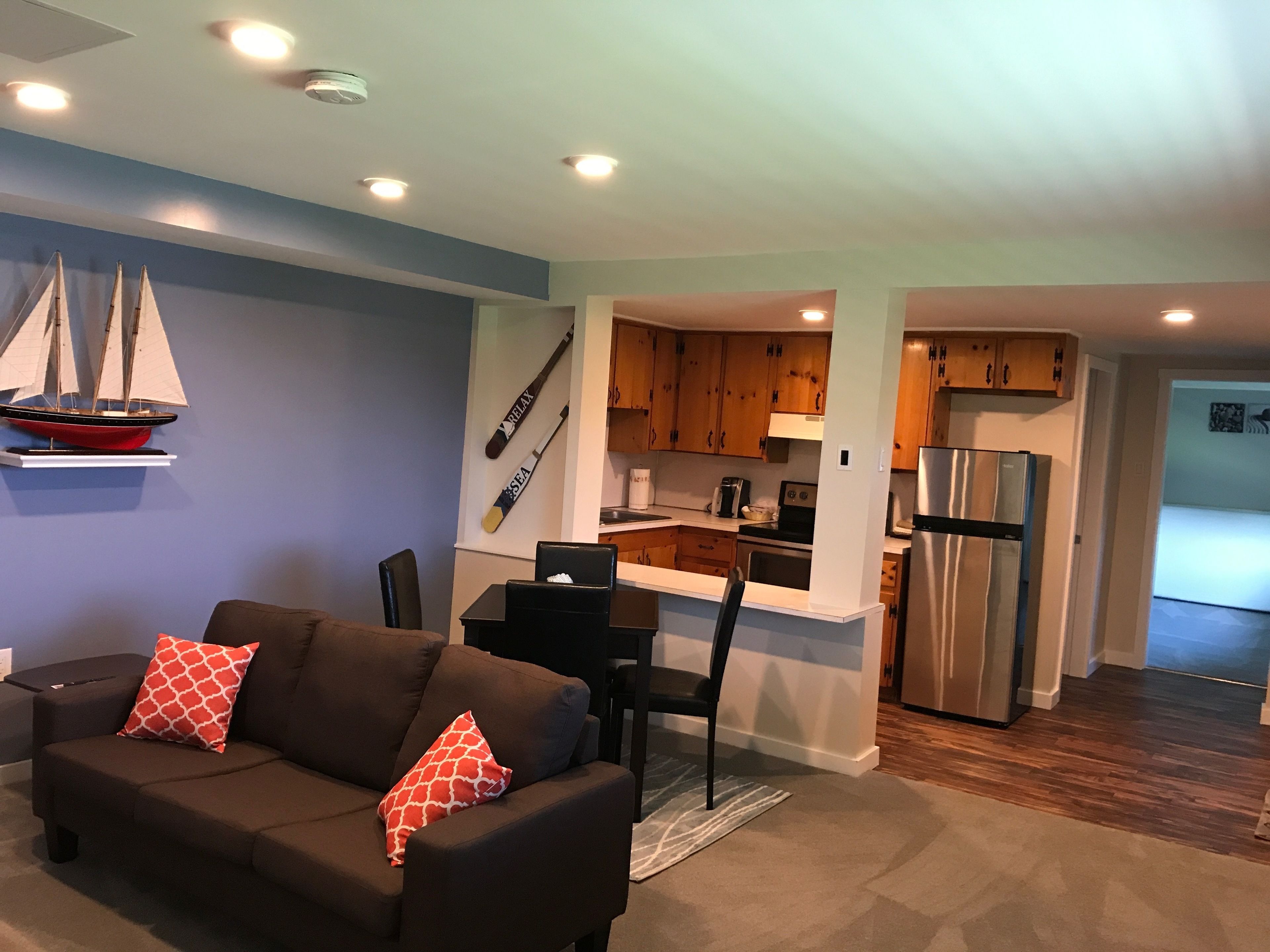 A modern living room with a dark brown sofa, a small table, and a red patterned pillow. A kitchen area is visible with wooden cabinets, a refrigerator, and a stove.