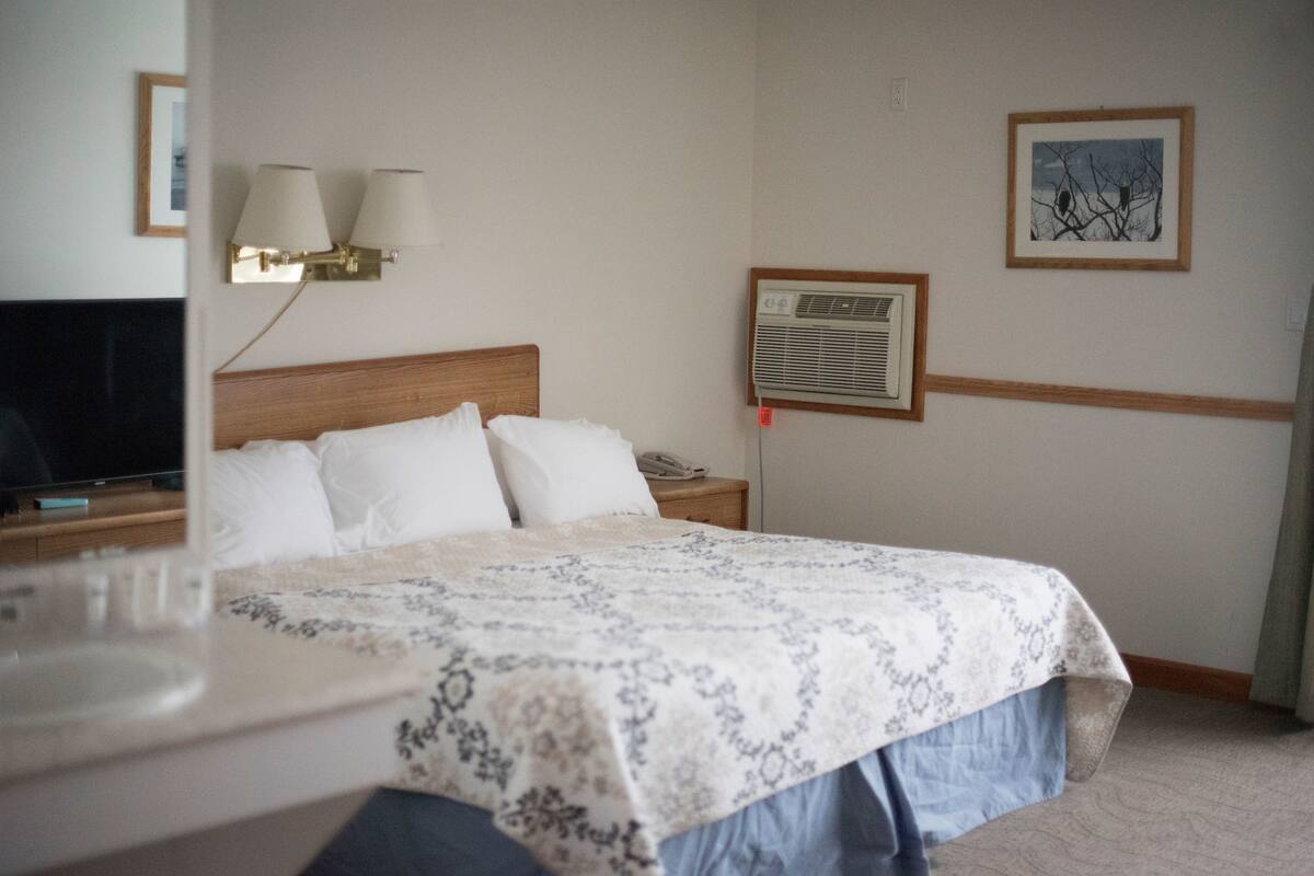 A hotel room with a bed, bedside lamps, an air conditioning unit, a framed picture on the wall, and a television on a stand.