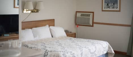 Una habitación de hotel con cama, lámparas de noche, aire acondicionado, un cuadro en la pared y televisor sobre un soporte.
