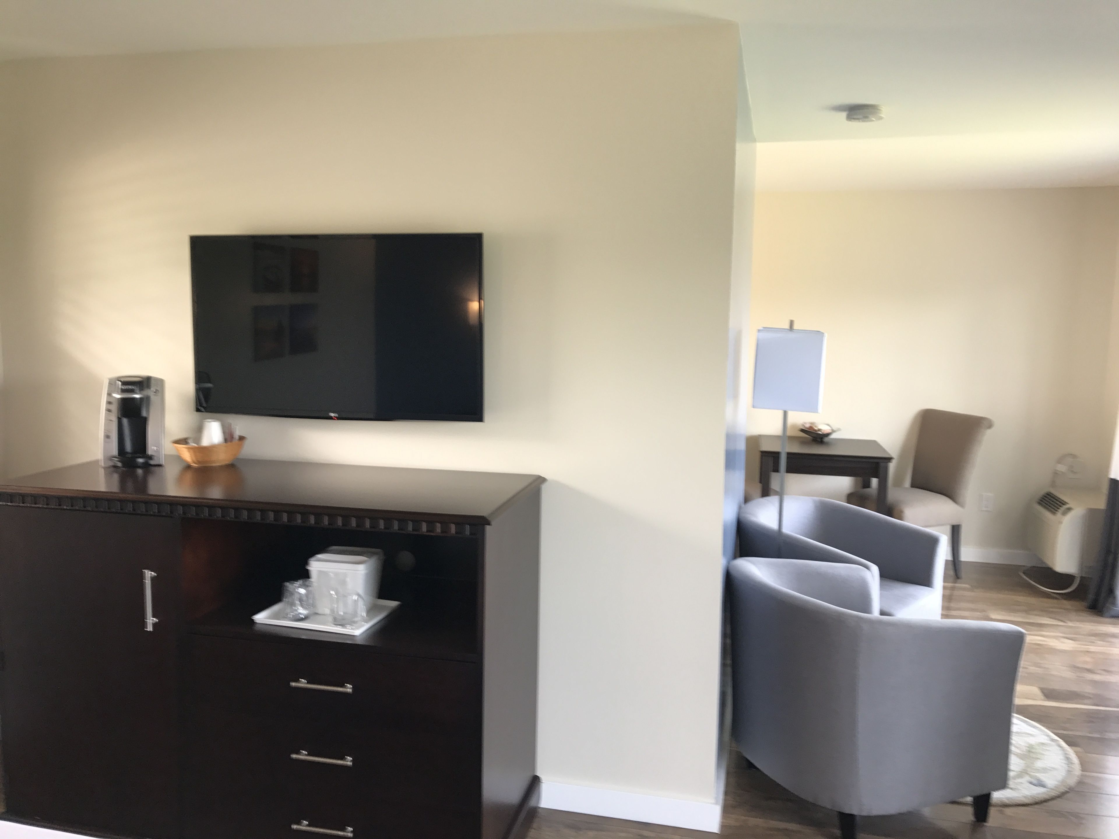 A hotel room with a dark wooden cabinet, a flat-screen TV mounted on the wall, a coffee maker, and a small table with a lamp and a bowl. There are two gray armchairs and a small table with a chair in the background.
