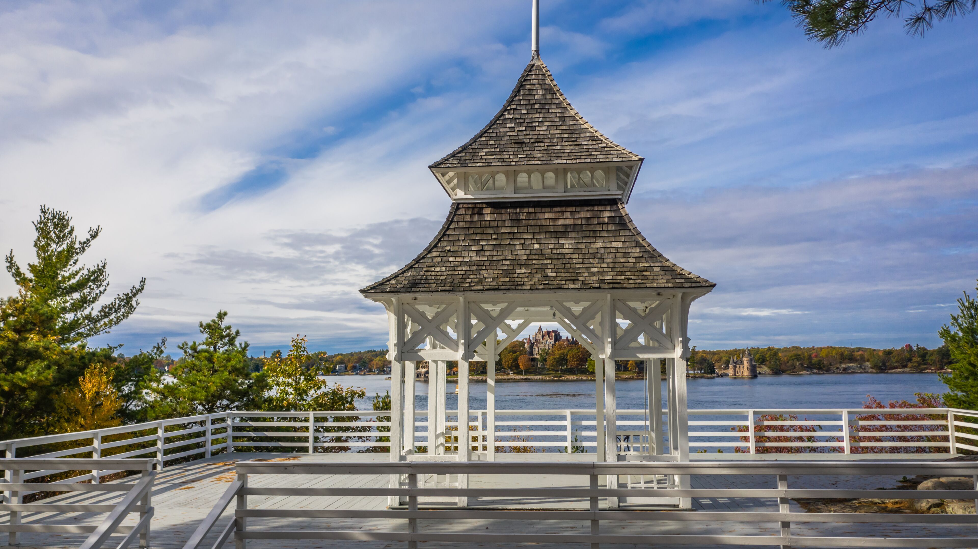 Ein Pavillon mit spitzer Kuppel, weißen Geländern und Blick auf eine Burg über dem Wasser.