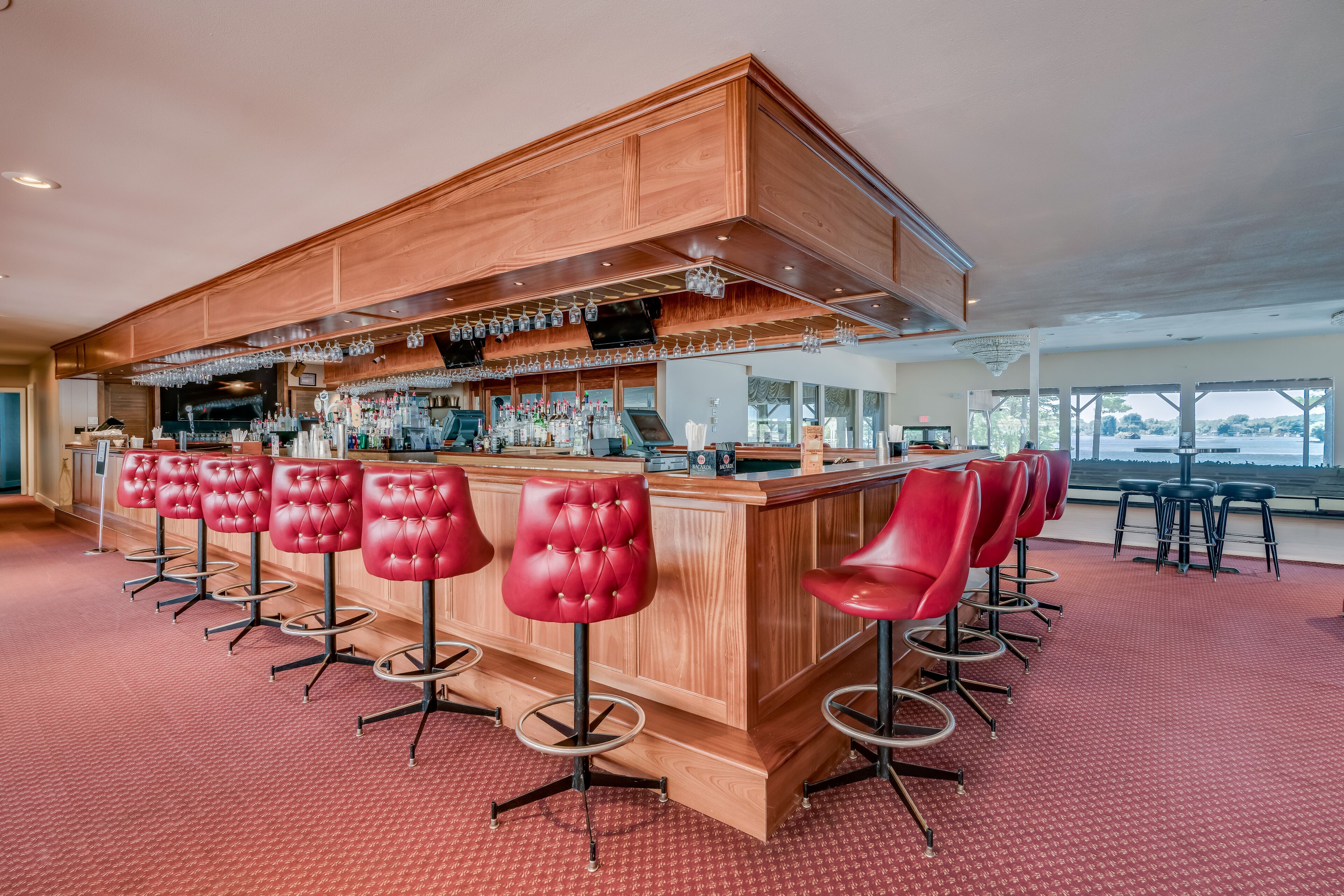 A bar area with wooden bar counter and red bar stools, a large window with a view, and a carpeted floor.