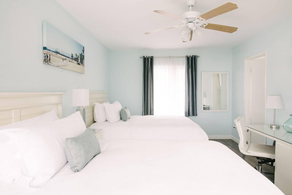 A hotel room with a large bed, a desk, a chair, a ceiling fan, a window with curtains, and a painting on the wall.