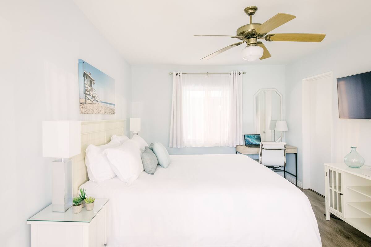 A bedroom with a large bed, a ceiling fan, a desk with a chair, a TV, and a window with curtains.