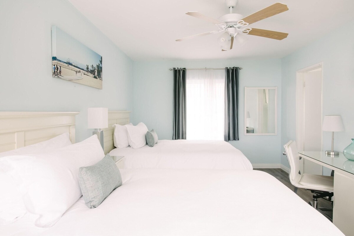 A hotel room with a large bed, a desk with a chair, a ceiling fan, a window with curtains, and a painting on the wall.