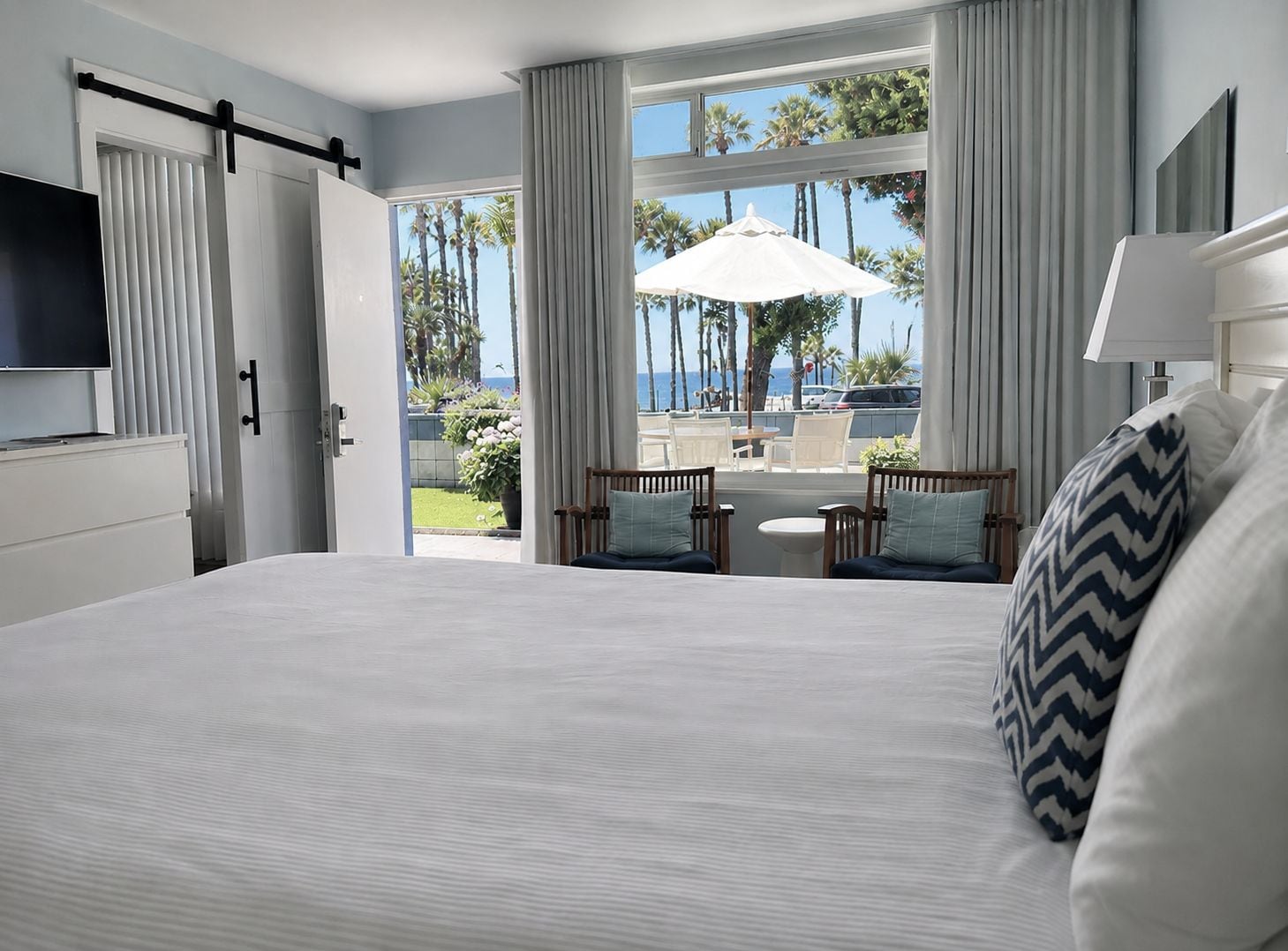 Habitación de hotel con una cama grande, televisión, balcón con mesa y sillas, y vistas a palmeras y al océano.