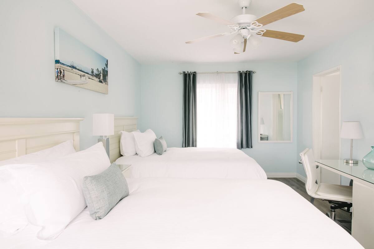 A hotel room with two beds, a ceiling fan, a desk with a chair, a mirror, and a painting on the wall.