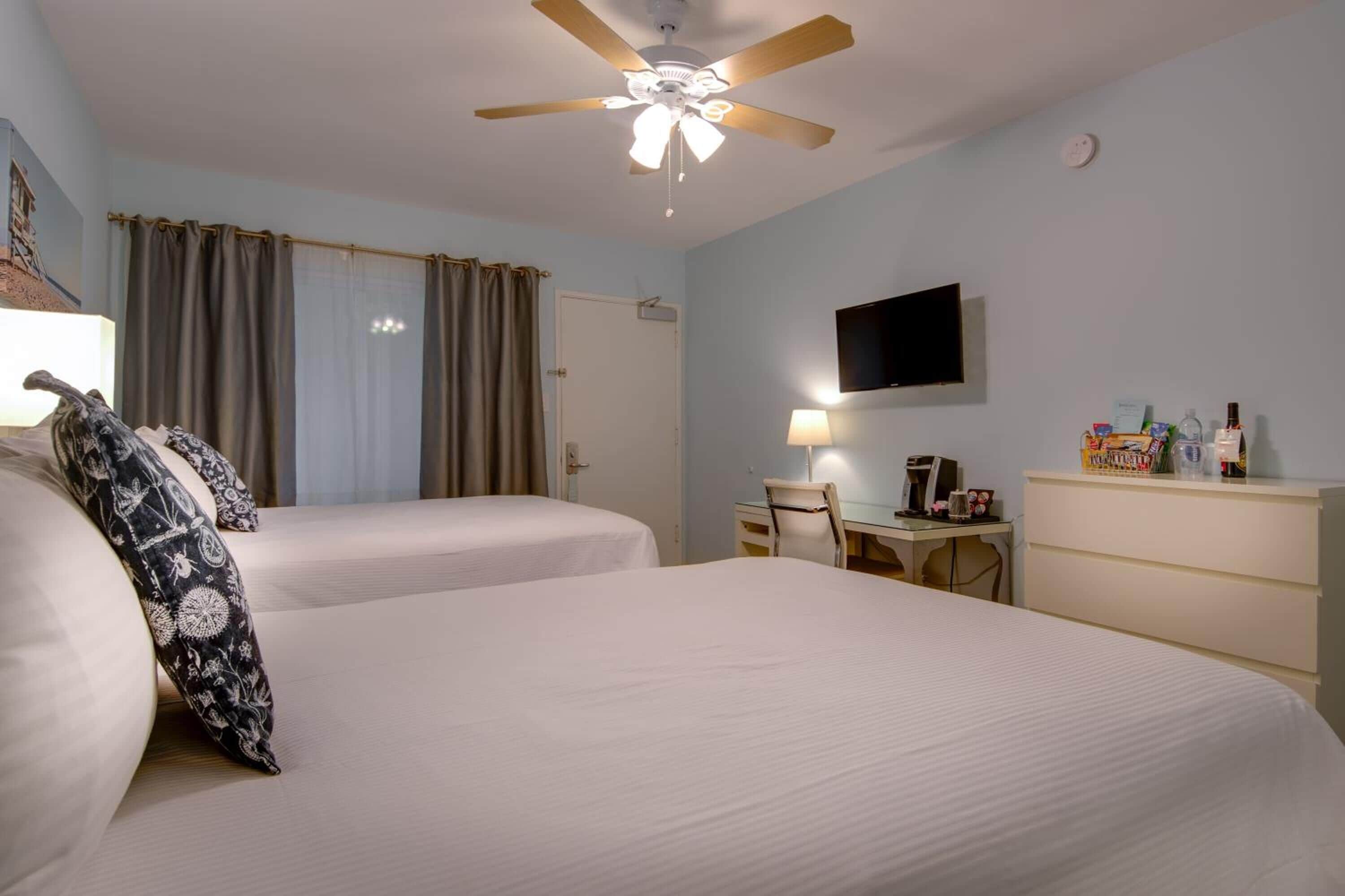 Ein Hotelzimmer mit zwei Betten, einem Schreibtisch mit Stuhl, einem Fernseher und einem Deckenventilator.