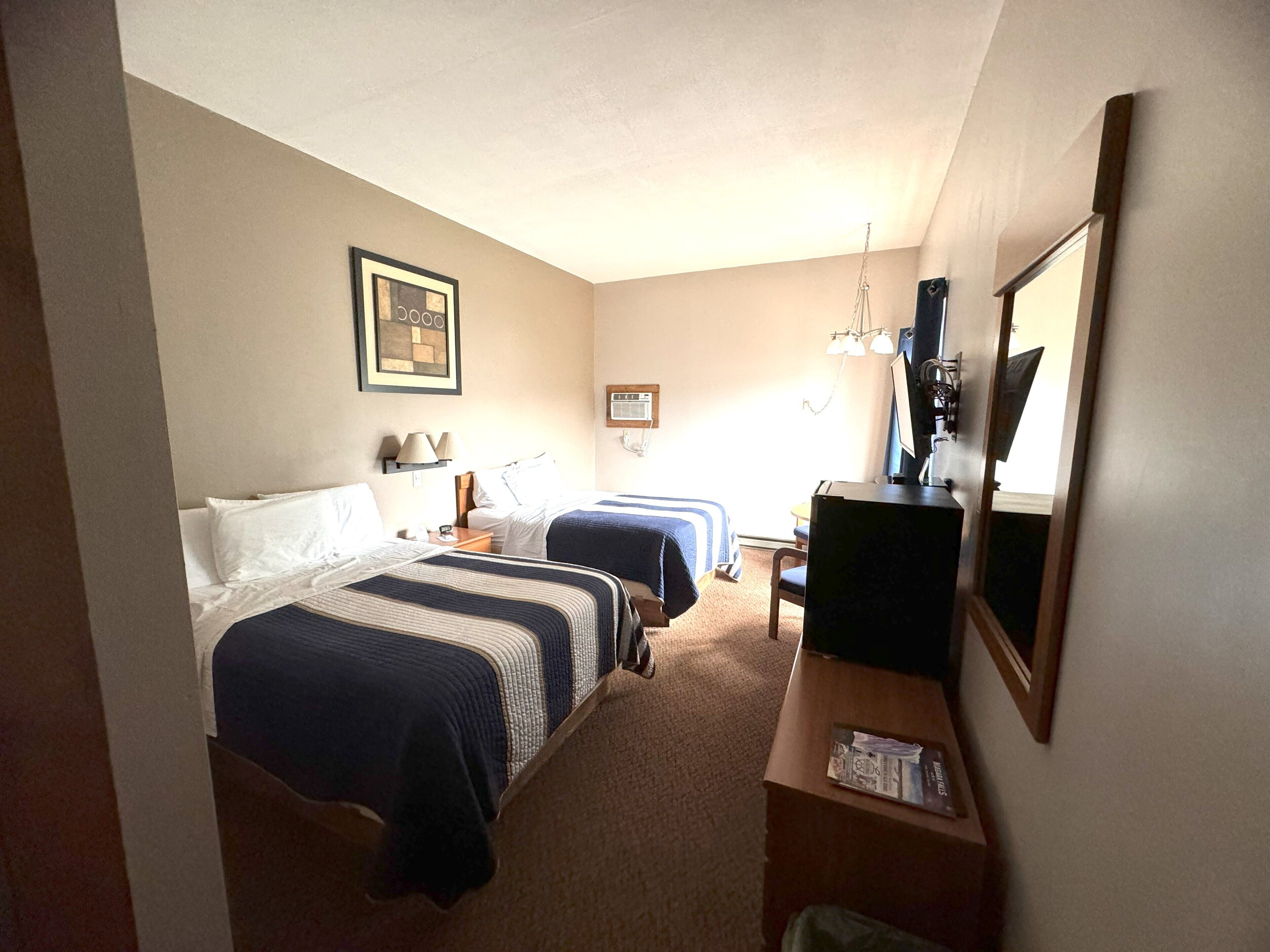 A hotel room with two beds, a desk, a chair, a desk lamp, a mirror, and a framed picture on the wall.