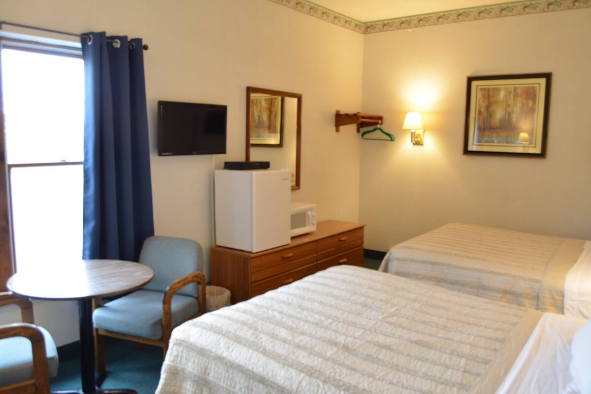 A hotel room with two beds, a small table with a chair, a microwave, a dresser, and a TV on the wall.