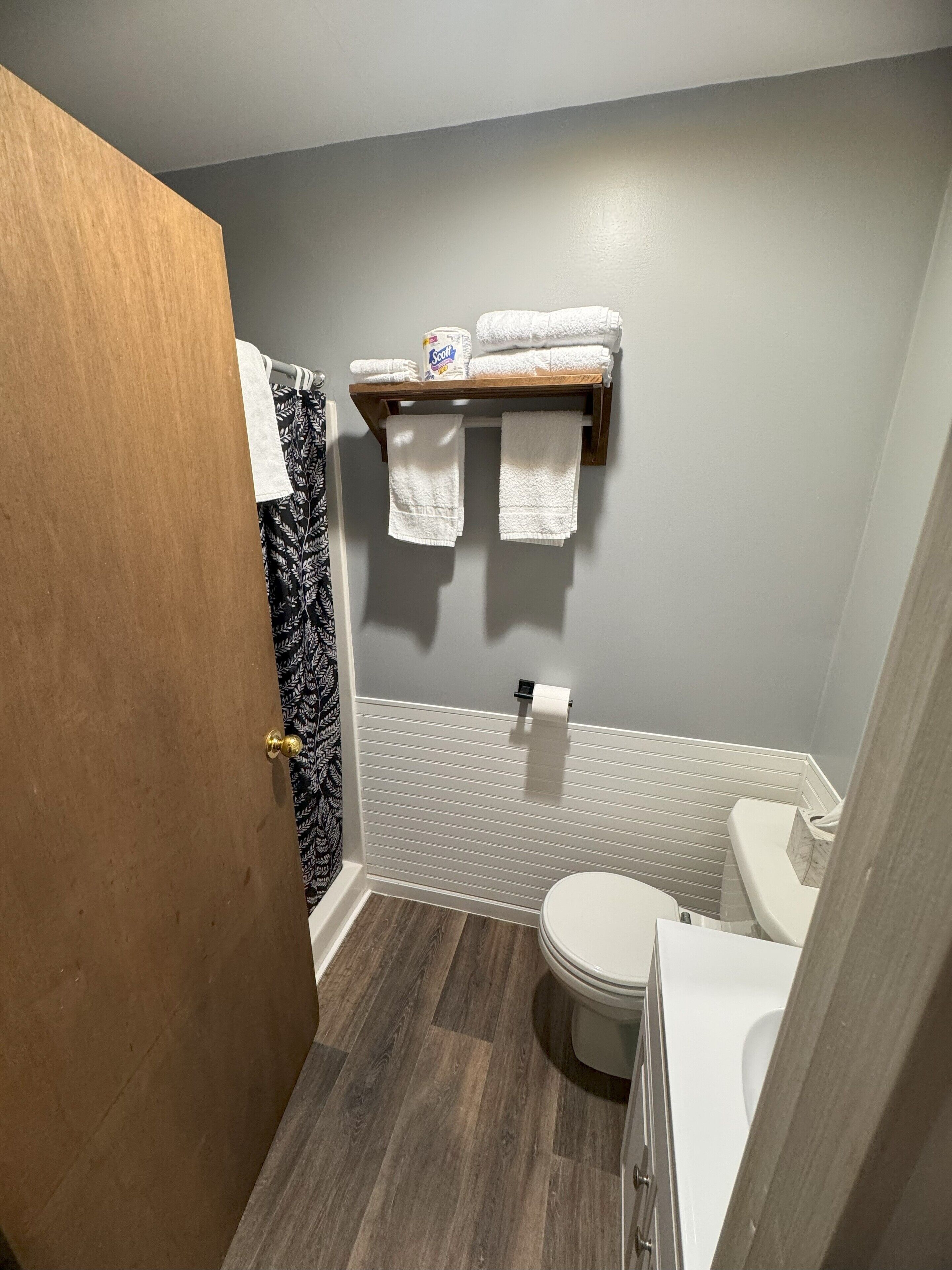 Une salle de bain avec une porte en bois, une douche dotée d’un rideau à motifs noir et blanc, des toilettes et une étagère avec des serviettes et un distributeur de papier toilette de la marque Scott.