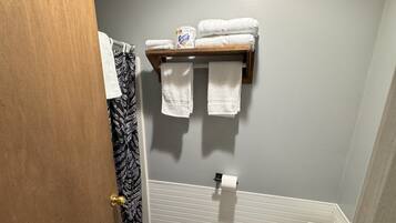 Une salle de bain avec une porte en bois, une douche dotée d’un rideau à motifs noir et blanc, des toilettes et une étagère avec des serviettes et un distributeur de papier toilette de la marque Scott.