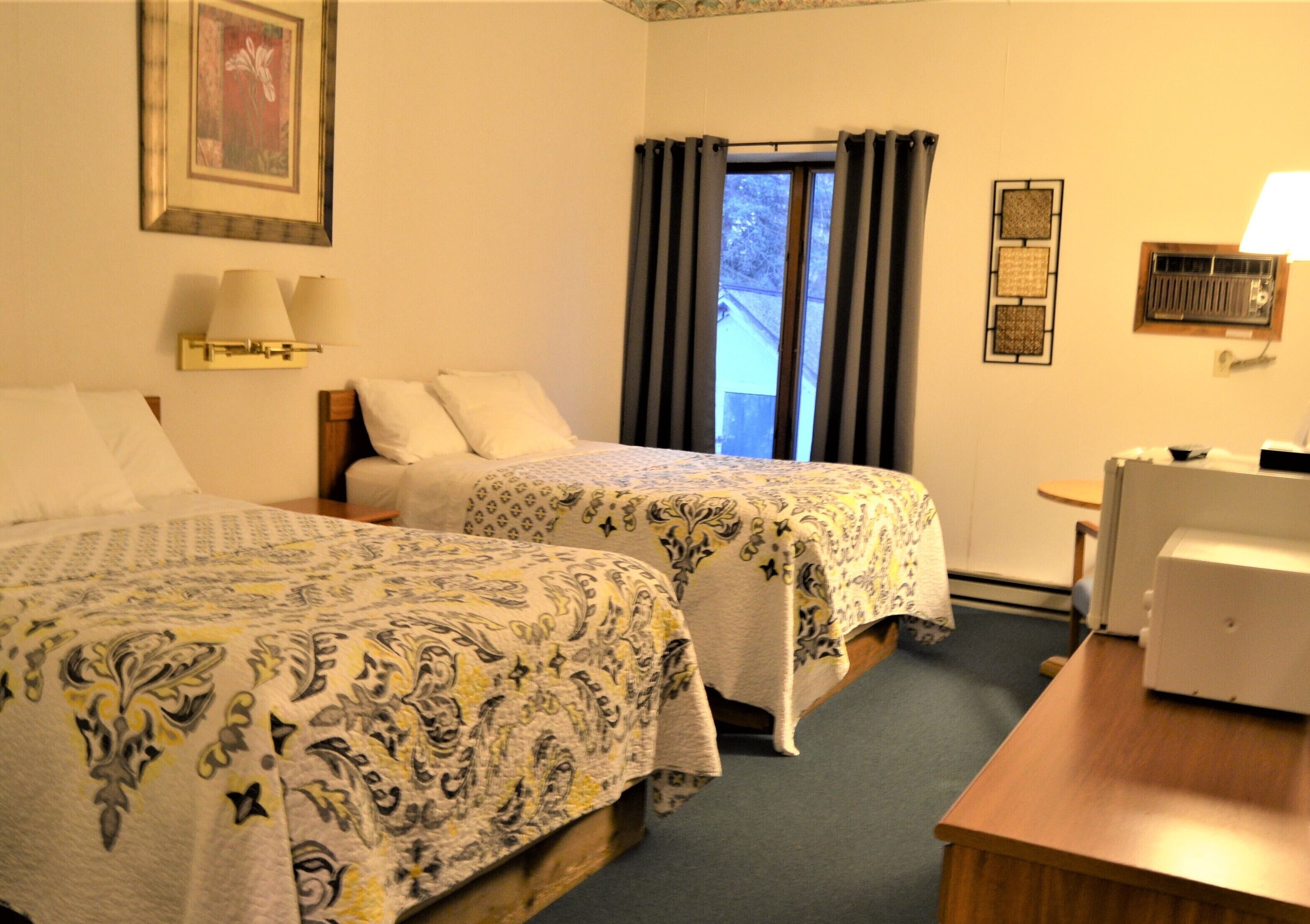 A hotel room with two beds, a desk, a chair, a window with curtains, a radiator, and wall art.