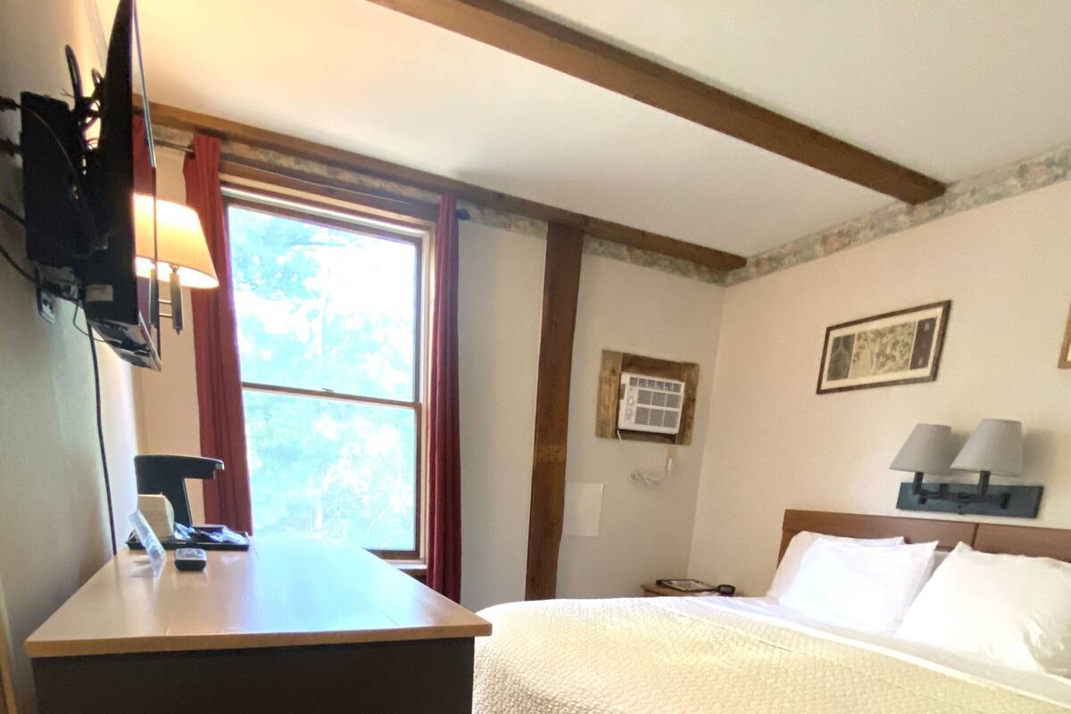 A hotel room with a bed, a desk, a window with curtains, a wall-mounted air conditioner, and a lamp.