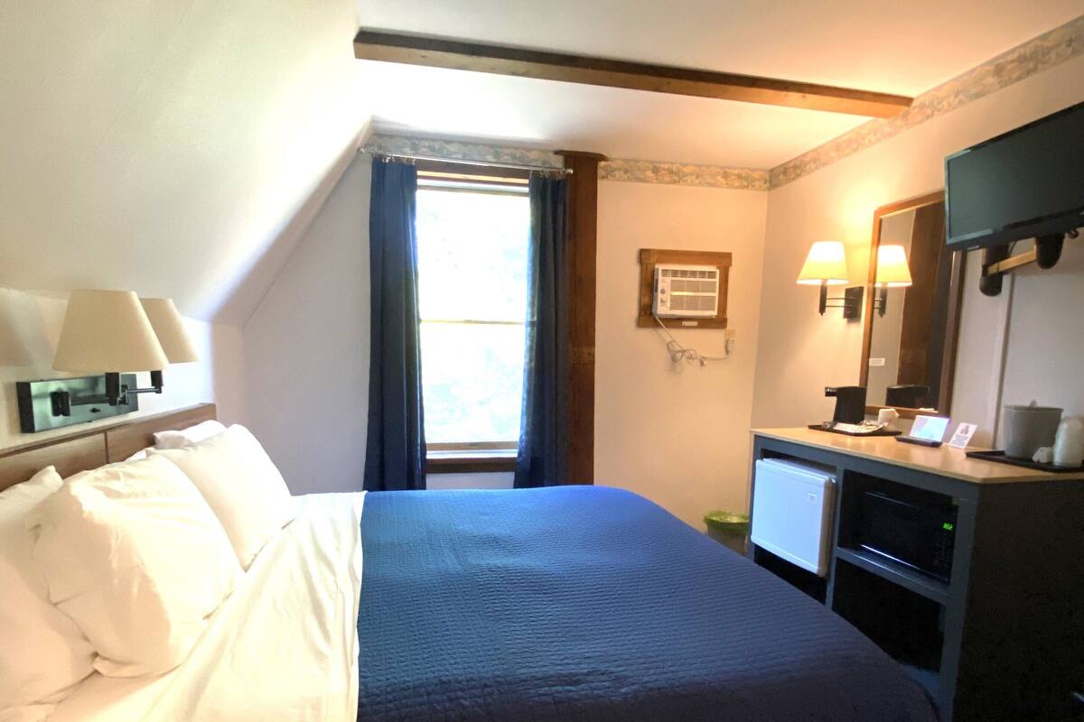 A hotel room with a bed, a desk with a computer, a mirror, and a window with curtains.