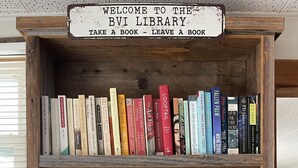 Una estantería de madera con un letrero que dice "BIENVENIDOS A LA BIBLIOTECA DE LAS VÍRGENES ISLASESCUCHO UN LIBRO - DEJA UN LIBRO".