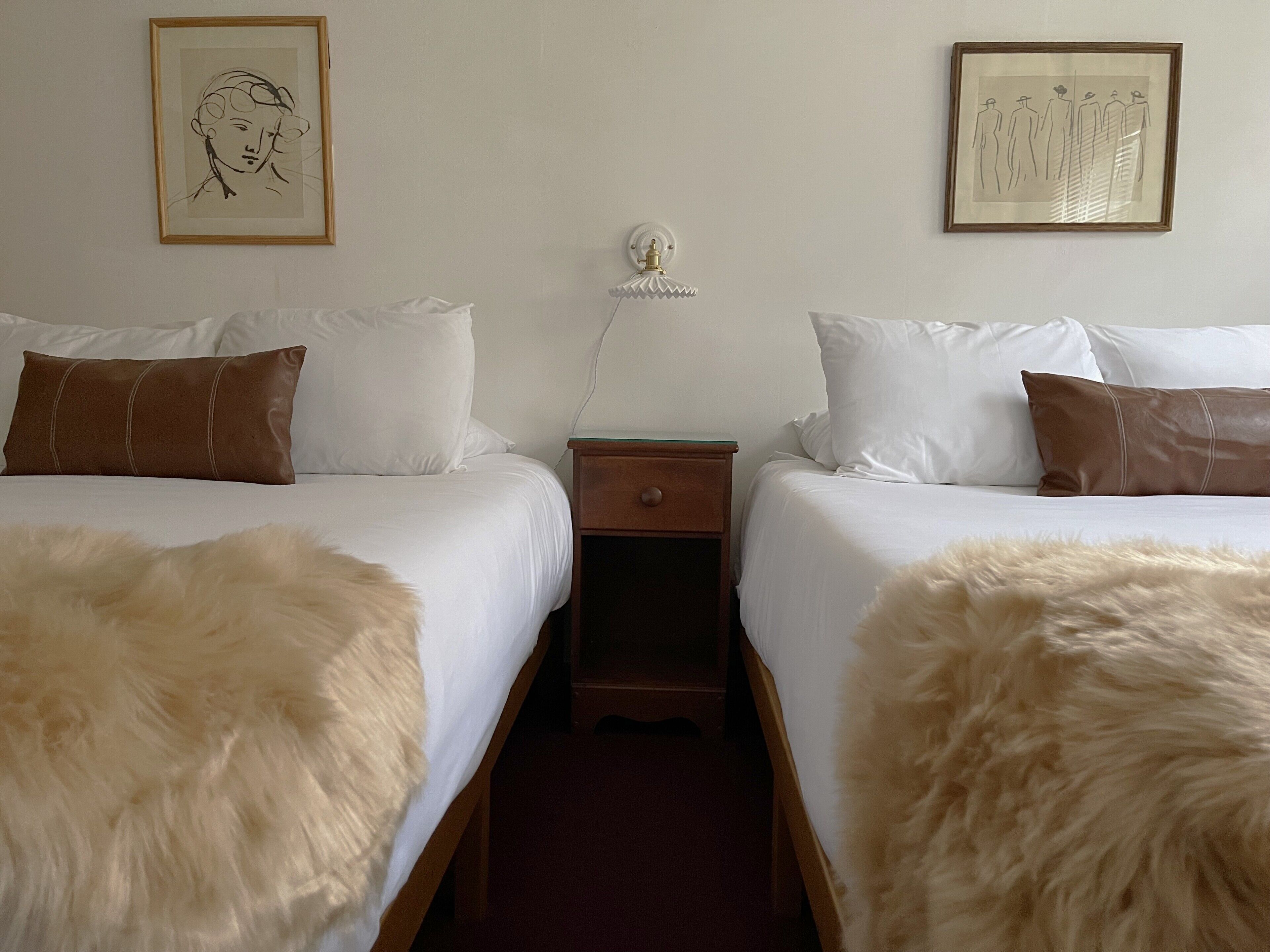 Habitación de hotel con dos camas, ropa de cama blanca, almohadas marrones, una mesita de noche de madera y una lámpara fijada a la pared.