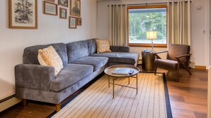 Una sala de estar con un sofá seccional gris, una pequeña mesa redonda de centro, un sillón de cuero marrón y una ventana con cortinas.