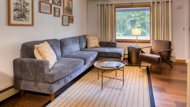 Una sala de estar con un sofá seccional gris, una pequeña mesa redonda de centro, un sillón de cuero marrón y una ventana con cortinas.