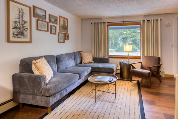 Una sala de estar con un sofá seccional gris, una pequeña mesa redonda de centro, un sillón de cuero marrón y una ventana con cortinas.
