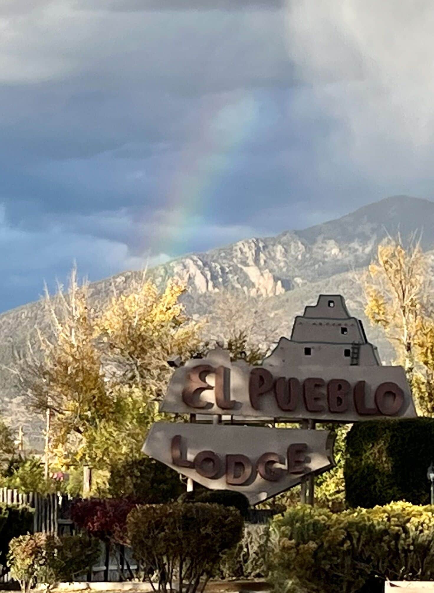 A sign for El Pueblo Lodge with a mountain backdrop and a rainbow.
