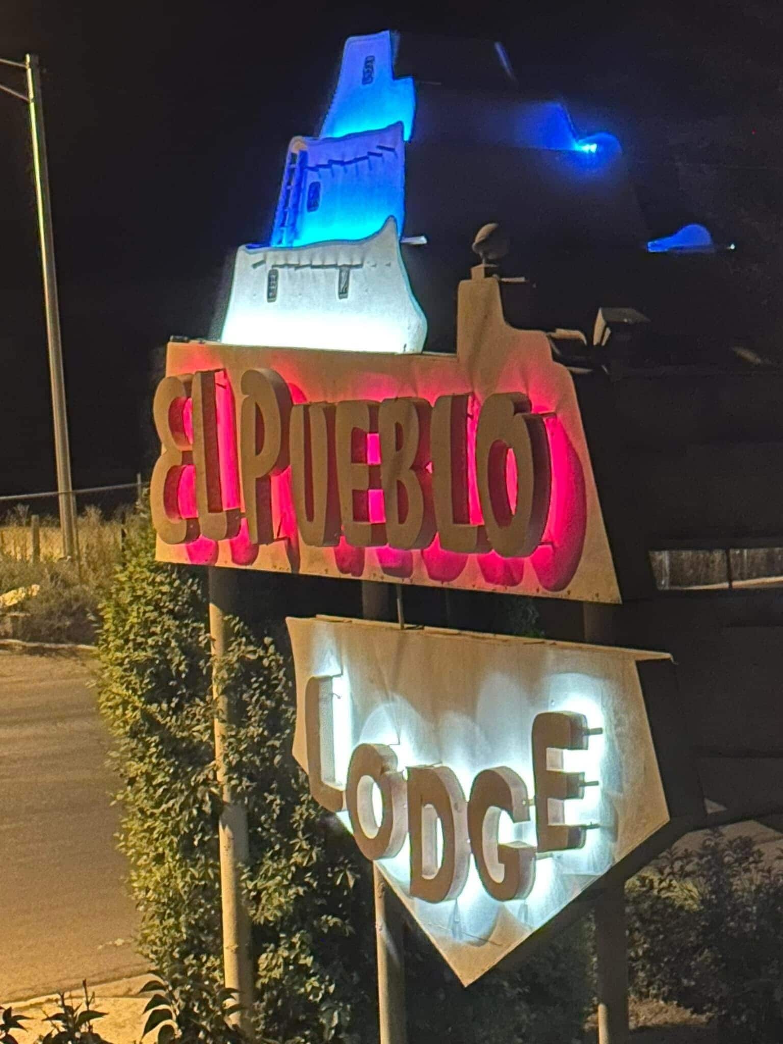 A sign for 'El Pueblo Lodge' with illuminated letters and a blue light on top.