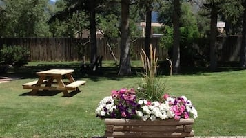 Uma mesa de piquenique e um vaso de flores num gramado com árvores e uma cerca ao fundo.