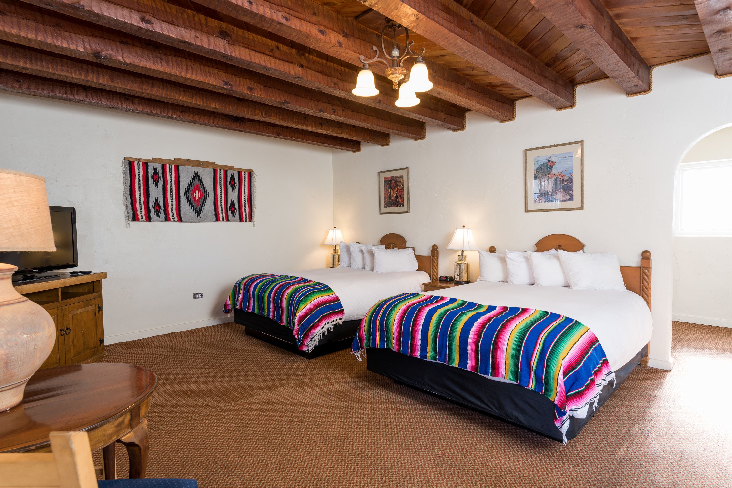 A hotel room with two beds, wooden beams on the ceiling, a television on a wooden cabinet, and artwork on the walls.