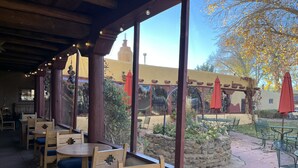An outdoor patio with wooden tables and chairs, a stone wall, and a view of a building with a red roof. - El Pueblo Lodge (Taos)