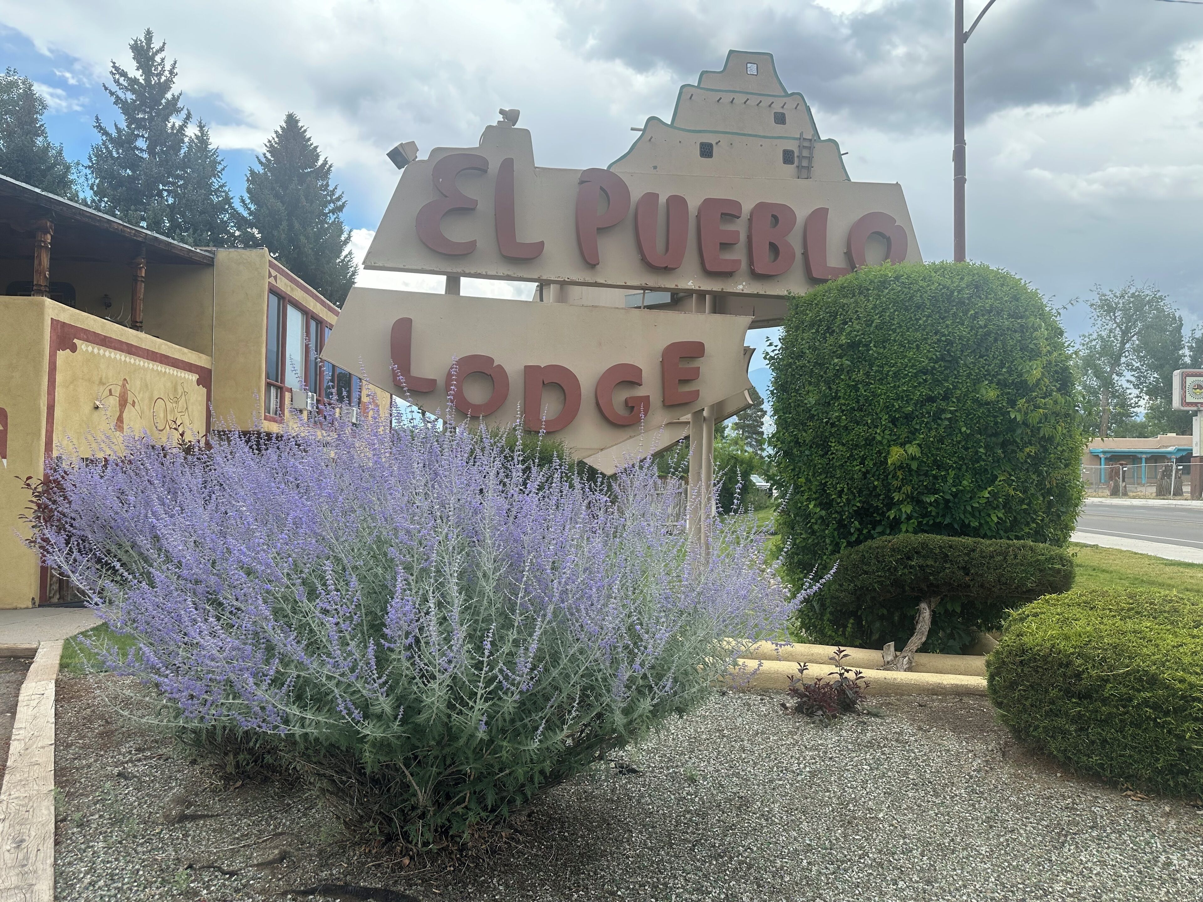 A sign for El Pueblo Lodge with a decorative design and a garden with purple flowers.