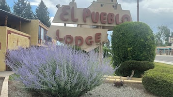 A sign for El Pueblo Lodge with a decorative design and a garden with purple flowers.