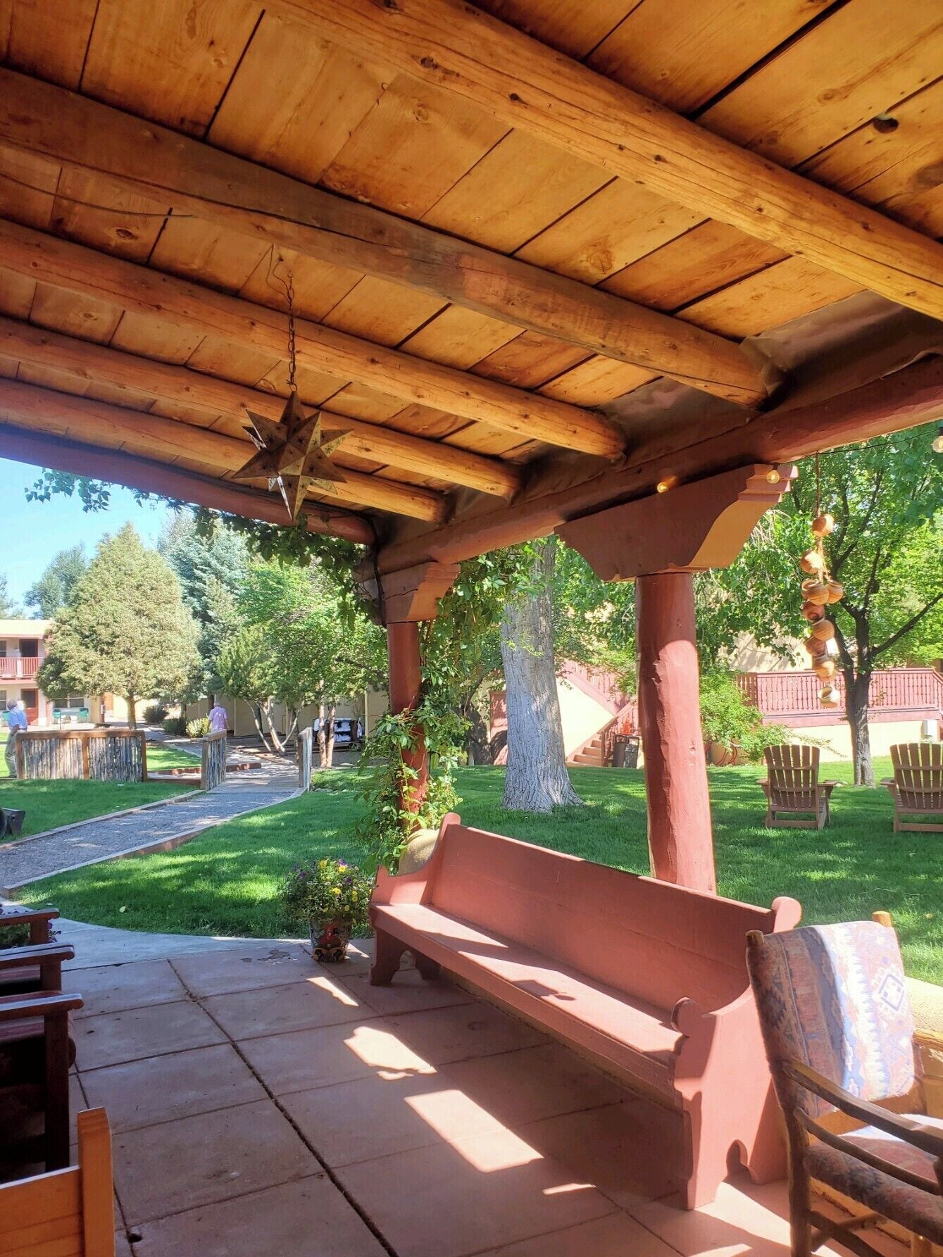 Un patio techado con un banco y sillas de madera, una lámpara colgante en forma de estrella, y vistas a un césped verde y árboles.